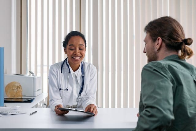 A smiling female doctor in a white lab coat with a stethoscope holding a tablet while consulting with a male patient at a desk