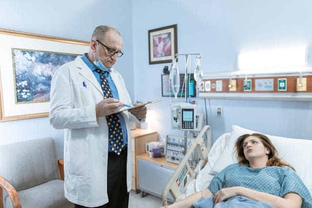 A male doctor in a white coat with a stethoscope and clipboard consulting with a female patient lying in a medical clinic bed