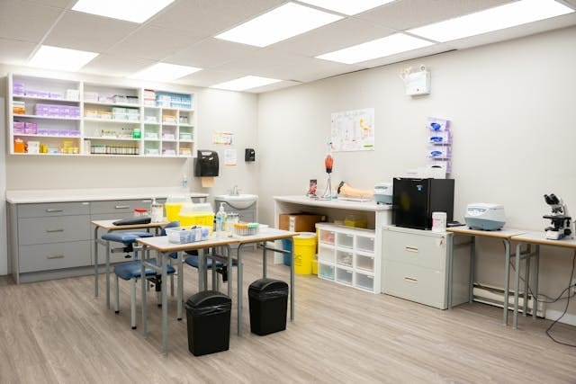 A clean, modern, and well-lit medical laboratory featuring clinical workstations, microscopes, and organized medical supply shelving