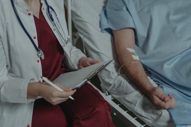 A medical professional in a lab coat with a stethoscope holding a clipboard while sitting next to a patient with an IV line in their arm