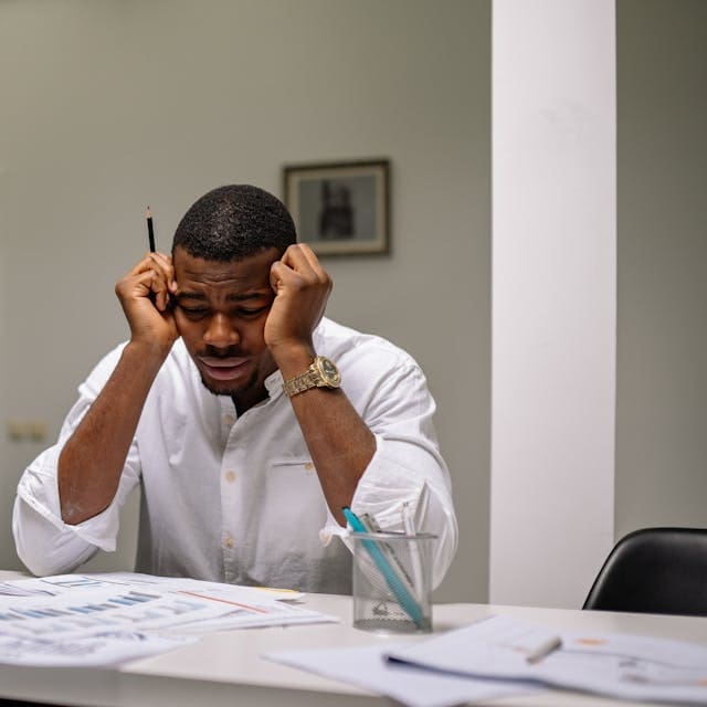 A stressed young man in a white shirt sitting at a desk with his head in his hands, looking down at paperwork and reports