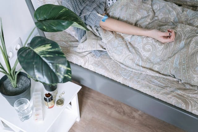 A patient resting in bed while receiving an intravenous (IV) infusion, with supplements, water, and a plant on a bedside table.