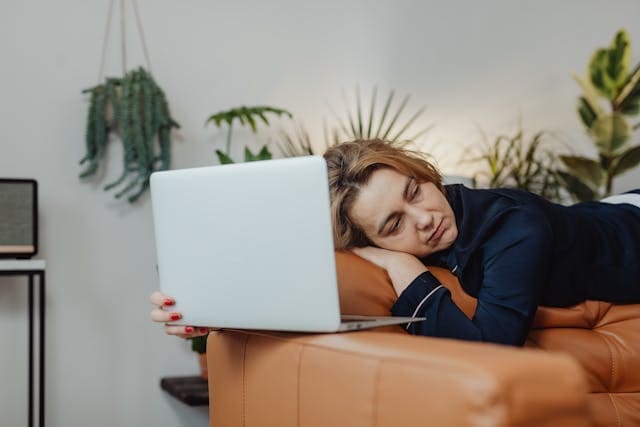 An exhausted woman lying on a brown leather couch while staring lethargically at a laptop, representing symptoms of persistent low energy and chronic fatigue