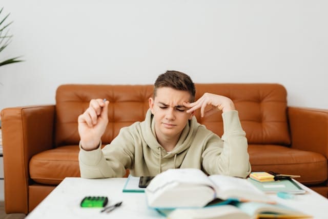 A young man appearing stressed and rubbing his temple while studying at a desk with books, representing high mental demands and fatigue