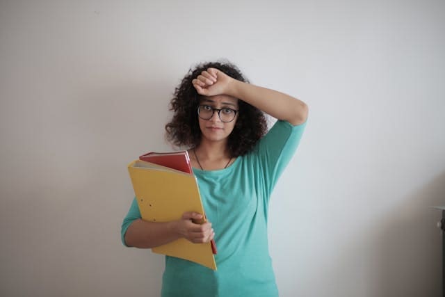 A woman in a green shirt holding work folders and resting her hand on her forehead, representing symptoms of mental fatigue and cognitive load.
